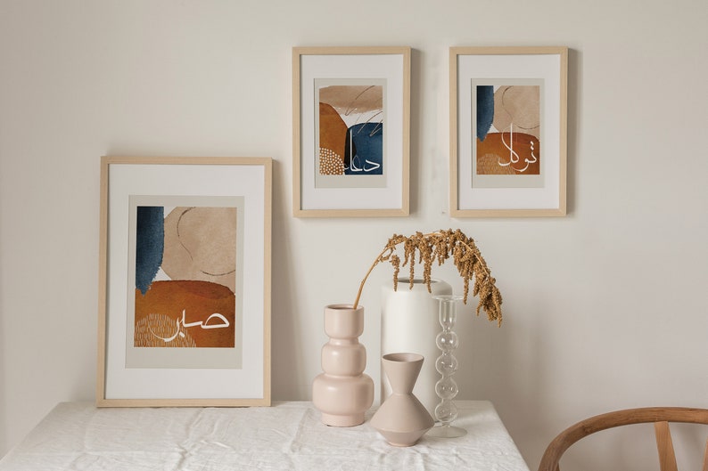 May include: Three framed abstract art prints featuring Arabic calligraphy and geometric shapes in shades of brown, beige, and blue. The prints are hanging on a white wall above a table with a vase of dried flowers.