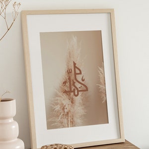 May include: A framed print with a beige background featuring a brown calligraphy design of the word "Jalla" in Arabic script. The design is centered in front of a single beige pampas grass plume.