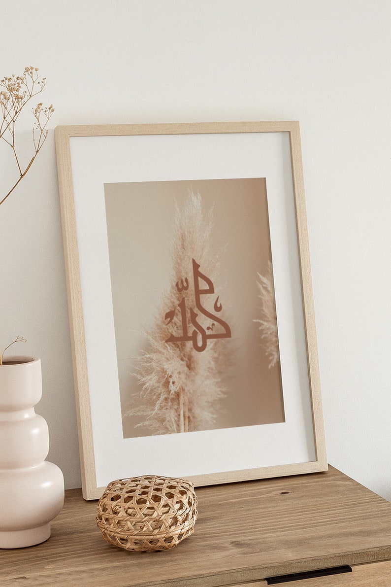 May include: A framed print with a beige background featuring a brown calligraphy design of the word "Jalla" in Arabic script. The design is centered in front of a single beige pampas grass plume.
