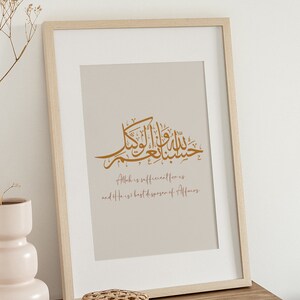 Op de afbeelding: Een ingelijst afdruk met een beige achtergrond met Arabische kalligrafie in goud. De tekst luidt "Allah is voldoende voor ons en Hij is de beste beschikker van zaken."