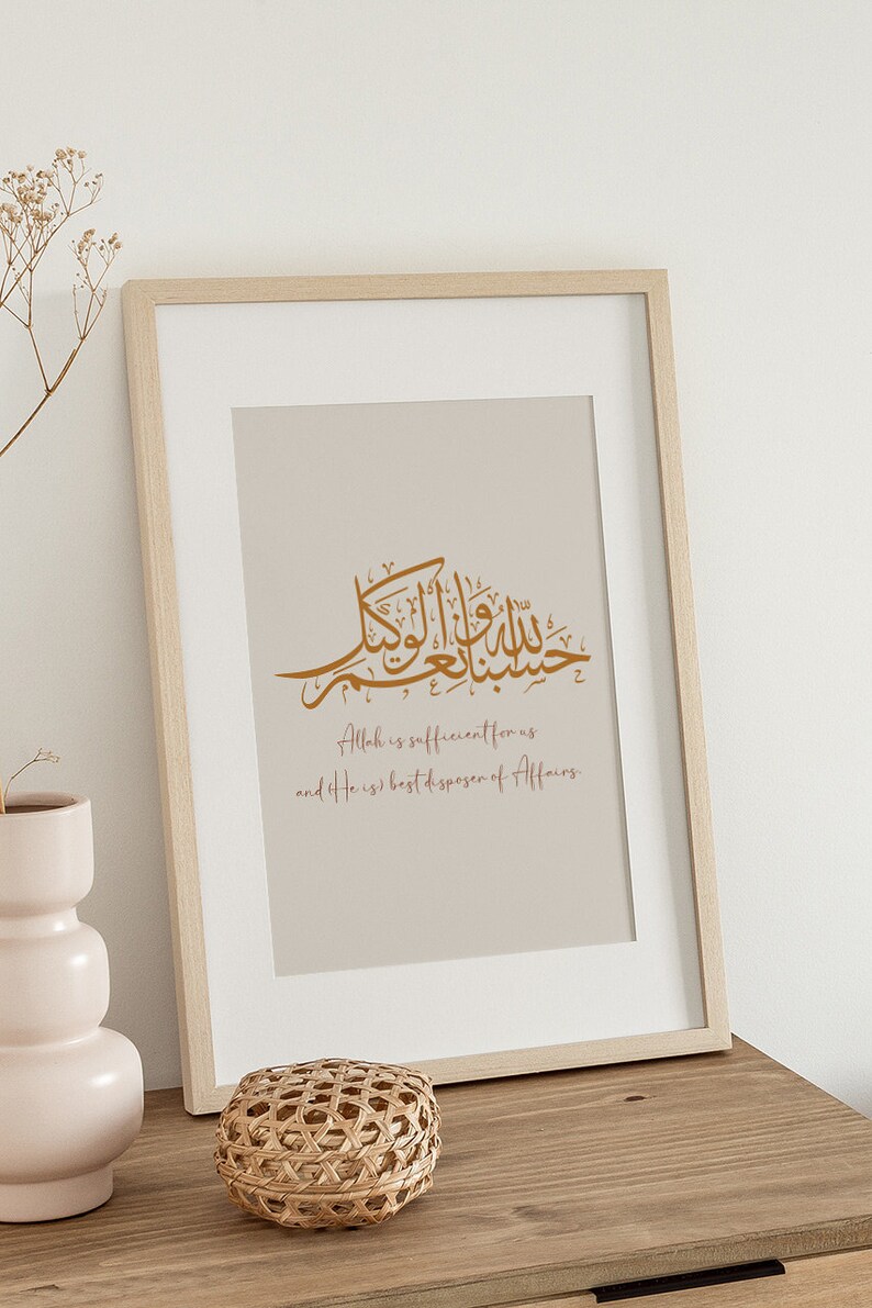 Op de afbeelding: Een ingelijst afdruk met een beige achtergrond met Arabische kalligrafie in goud. De tekst luidt "Allah is voldoende voor ons en Hij is de beste beschikker van zaken."