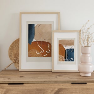 May include: Two framed abstract art prints with a beige background and blue and orange shapes. The prints feature Arabic calligraphy in white. The prints are hanging on a light wood wall shelf.