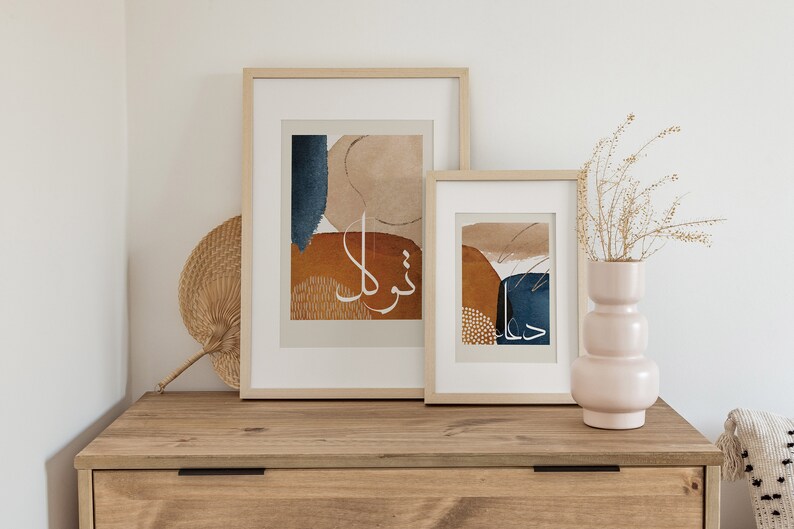 May include: Two framed abstract art prints with a beige background and blue and orange shapes. The prints feature Arabic calligraphy in white. The prints are hanging on a light wood wall shelf.
