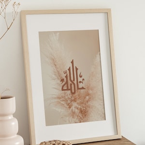 May include: A framed print with a beige background featuring the Arabic word "Allah" in brown calligraphy. The word is centered on a single, dried pampas grass plume.