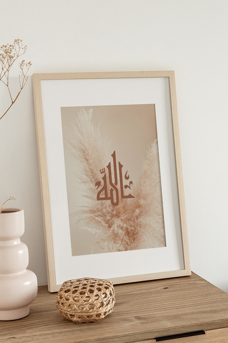 May include: A framed print with a beige background featuring the Arabic word "Allah" in brown calligraphy. The word is centered on a single, dried pampas grass plume.