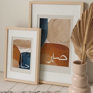May include: Two framed abstract art prints with a beige background and blue, brown, and white abstract shapes. The larger print has the Arabic word "حياة" (meaning "life") written in white. The smaller print has the Arabic word "جمال" (meaning "beauty") written in white. The prints are displayed on a white surface with a beige rug and a beige vase with a dried palm leaf.