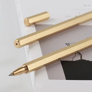 Puede incluir: Dos bolígrafos hexagonales dorados, uno con tapa y otro con una punta de tinta negra. Los bolígrafos están sobre una superficie blanca con una página de revista en el fondo.