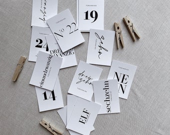 Może przedstawiać: Zbiór białych kart z czarnymi numerami i tekstem, w tym "19", "22", "24", "14", "ELF" i "sechzehn". Karty są rozrzucone na neutralnej powierzchni, z drewnianymi spinaczami. Karty są przeznaczone do kalendarza adwentowego.