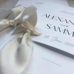May include: White wedding invitation with black text that reads "Einladung zur Hochzeit" and "Alexan and Samm" with the date "18. June 2024". The invitation is decorated with a white satin ribbon.