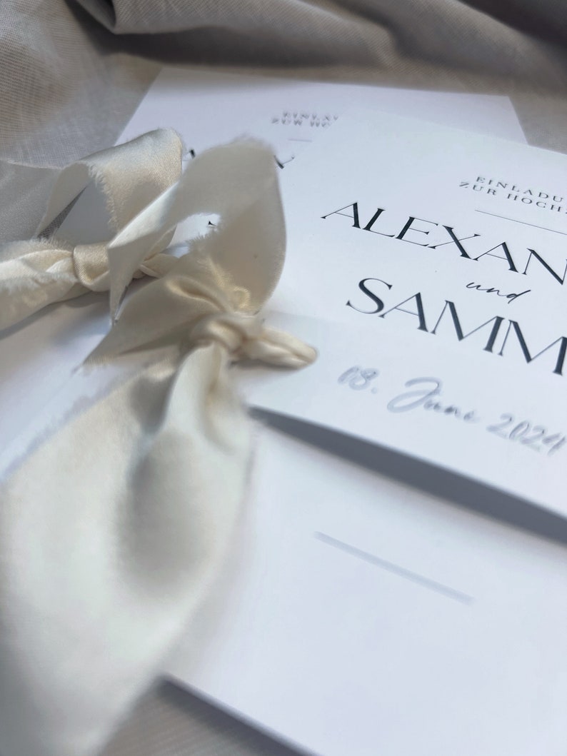 May include: White wedding invitation with black text that reads "Einladung zur Hochzeit" and "Alexan and Samm" with the date "18. June 2024". The invitation is decorated with a white satin ribbon.