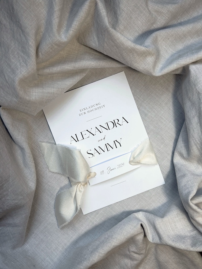 May include: A white wedding invitation with a white ribbon tied around it. The invitation reads "Einladung zur Hochzeit" and features the names "Alexandra and Sammy" and the date "08. Juni 2024".