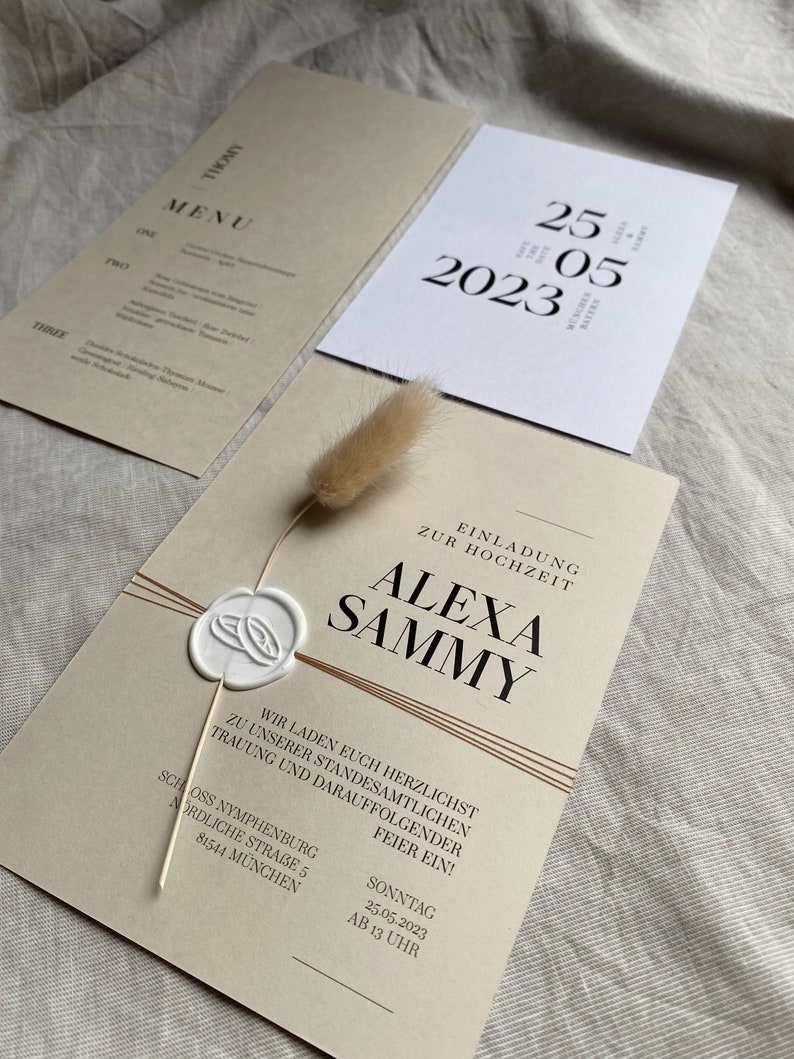May include: A wedding invitation with a beige background and brown accents. The invitation features the names "Alexa" and "Sammy" in a bold, modern font. The text reads "We invite you to our wedding ceremony and reception on Sunday, May 25, 2023 at 1 p.m. at Schloss Nymphenburg, Nordliche Stra&szlig;e 5, 81544 M&uuml;nchen."
