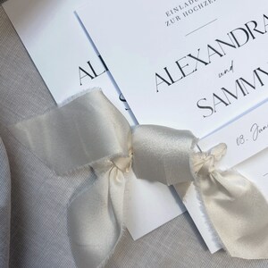 May include: Two white wedding invitations with a simple, modern design. The top invitation reads "Einladung zur Hochzeit" and the bottom invitation reads "Alexandra and Sammy". The invitations are tied together with a white satin ribbon.