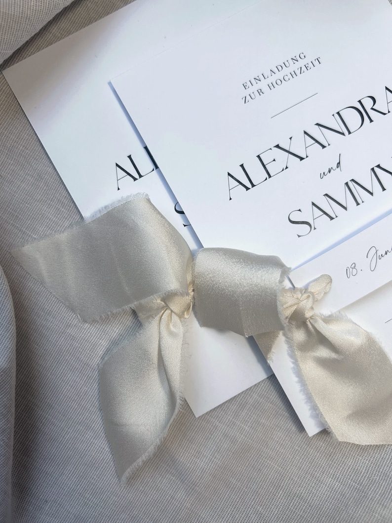 May include: Two white wedding invitations with a simple, modern design. The top invitation reads "Einladung zur Hochzeit" and the bottom invitation reads "Alexandra and Sammy". The invitations are tied together with a white satin ribbon.