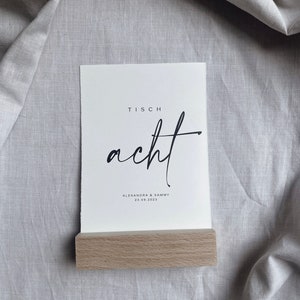 K&ouml;nnte beinhalten: Eine wei&szlig;e Karte mit dem Text "Tisch acht" in schwarzer Kalligraphie. Unter dem Text steht "Alexandra & Sammy 23.09.2023" in kleinerer schwarzer Schrift. Die Karte steht auf einem Holzst&auml;nder.