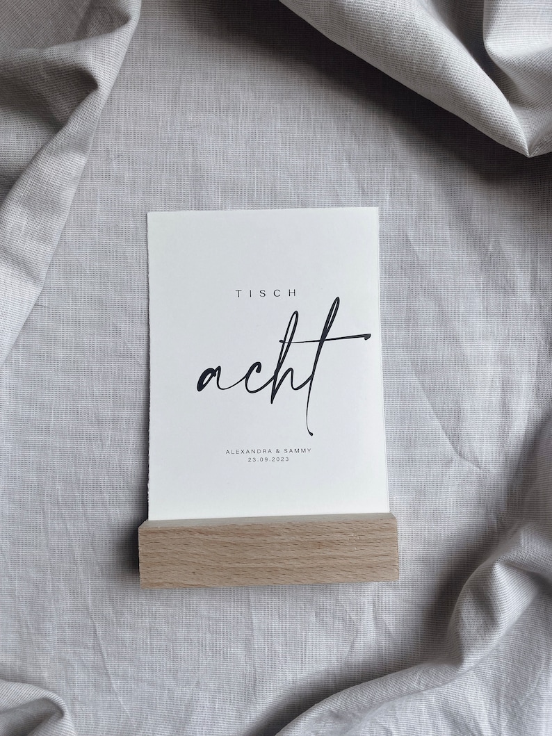 K&ouml;nnte beinhalten: Eine wei&szlig;e Karte mit dem Text "Tisch acht" in schwarzer Kalligraphie. Unter dem Text steht "Alexandra & Sammy 23.09.2023" in kleinerer schwarzer Schrift. Die Karte steht auf einem Holzst&auml;nder.