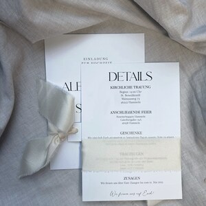 May include: A white wedding invitation with the text "Einladung zur Hochzeit" (Invitation to the wedding) and details about the ceremony and reception. The invitation is decorated with a white ribbon.