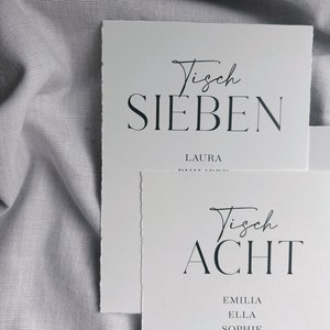 May include: Two white cards with black text. The top card says "Tisch Sieben" and "Laura". The bottom card says "Tisch Acht" and lists the names "Emilia", "Ella", "Sophie", "Mats", "Aleyna", "Amira".