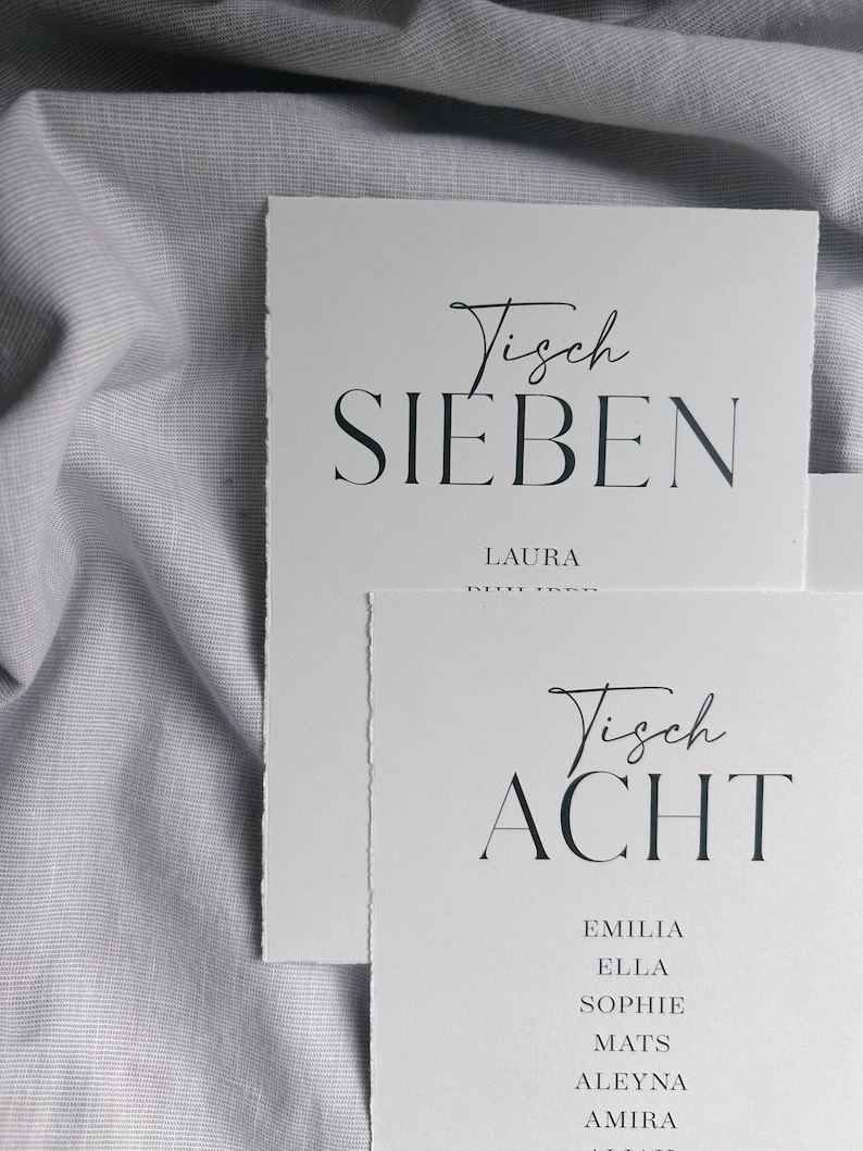 May include: Two white cards with black text. The top card says "Tisch Sieben" and "Laura". The bottom card says "Tisch Acht" and lists the names "Emilia", "Ella", "Sophie", "Mats", "Aleyna", "Amira".