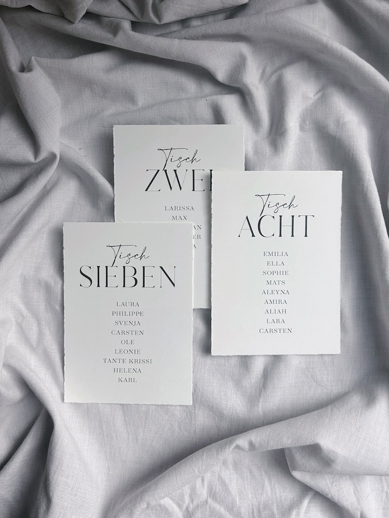 May include: Three white cards with black text, each labelled with a table number and a list of names. The cards are labelled "Tisch Zwei", "Tisch Sieben", and "Tisch Acht".
