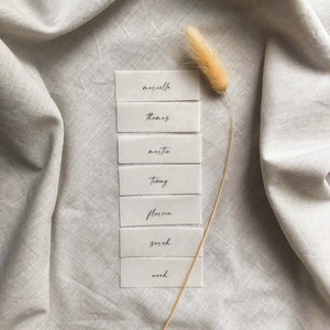 Puede incluir: Siete tarjetas de lugar rectangulares blancas con nombres en caligraf&iacute;a negra: Marcella, Thomas, Martin, Timmy, Florin, Sarah, Noah. Las tarjetas est&aacute;n dispuestas verticalmente sobre una superficie de tela gris claro con un tallo de hierba seca.