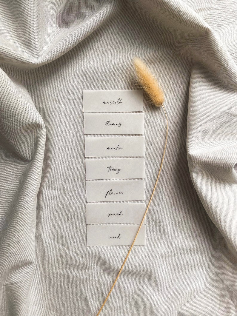 Puede incluir: Siete tarjetas de lugar rectangulares blancas con nombres en caligraf&iacute;a negra: Marcella, Thomas, Martin, Timmy, Florin, Sarah, Noah. Las tarjetas est&aacute;n dispuestas verticalmente sobre una superficie de tela gris claro con un tallo de hierba seca.