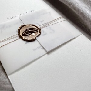 Op de afbeelding: Een witte kaart met de tekst "Save the Date" en een waszegel met twee gouden ringen. De kaart is vastgebonden met een bruin touwtje.