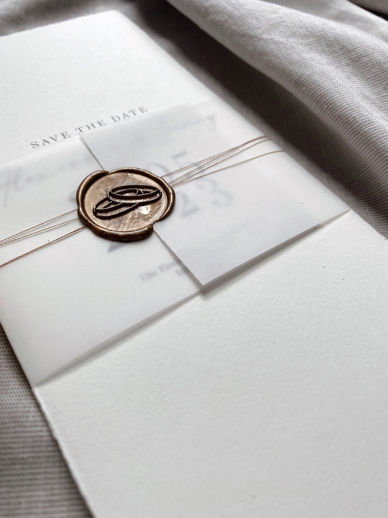 Op de afbeelding: Een witte kaart met de tekst "Save the Date" en een waszegel met twee gouden ringen. De kaart is vastgebonden met een bruin touwtje.