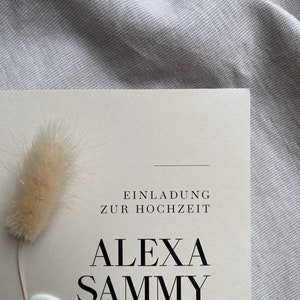 May include: A wedding invitation with a cream background and black text. The text reads "Einladung zur Hochzeit" and "Alexa Sammy". A white wax seal with two rings is in the bottom left corner.