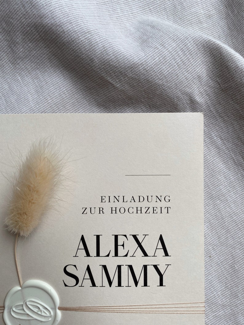 May include: A wedding invitation with a cream background and black text. The text reads "Einladung zur Hochzeit" and "Alexa Sammy". A white wax seal with two rings is in the bottom left corner.