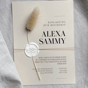 May include: A beige wedding invitation with a wax seal and a dried flower. The invitation reads "Einladung zur Hochzeit Alexa Sammy" and includes the date and time of the wedding: "Sonntag 25.05.2023 AB 13 UHR". The wedding will take place at Schloss Nymphenburg, Nordliche Strasse 5, 81544 M&uuml;nchen.