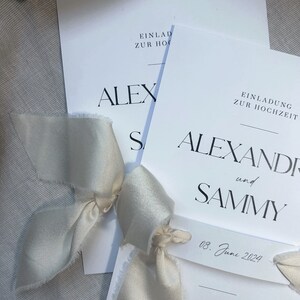 May include: Two white wedding invitations with black text. The top invitation reads "Einlad zur Hoch" and "ALEX". The bottom invitation reads "Einladung zur Hochzeit" and "ALEXANDER und SAMMY". The date "08. June 2024" is written on a small tag.