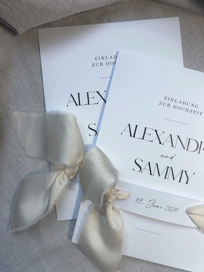 May include: Two white wedding invitations with black text. The top invitation reads "Einlad zur Hoch" and "ALEX". The bottom invitation reads "Einladung zur Hochzeit" and "ALEXANDER und SAMMY". The date "08. June 2024" is written on a small tag.