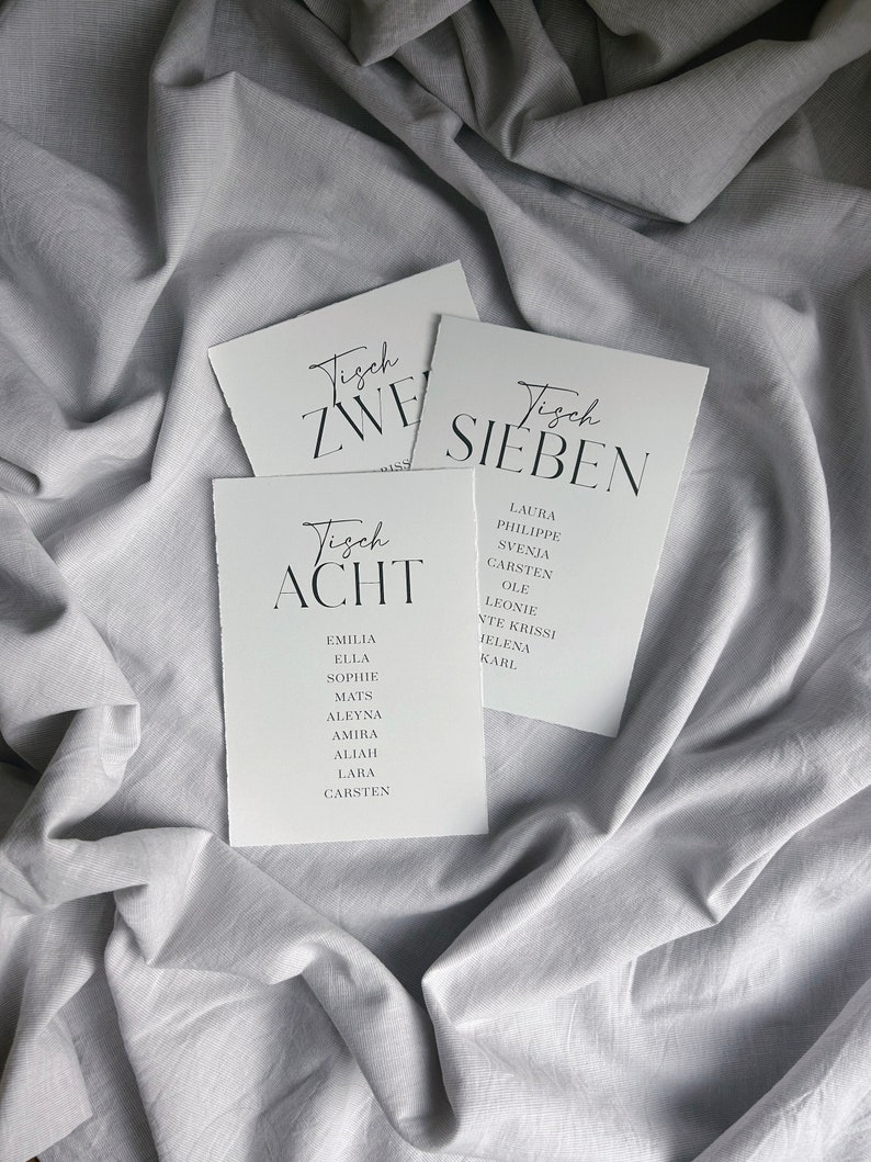 May include: Three white cards with black text. The cards are labelled "Tisch Zwei", "Tisch Sieben", and "Tisch Acht". Each card lists a group of names.