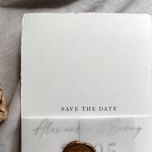 Op de afbeelding: Een witte save-the-date kaart met een gouden waszegel en de tekst "Save the Date" gedrukt op de voorkant. De kaart is vastgebonden met een dun bruin touwtje.