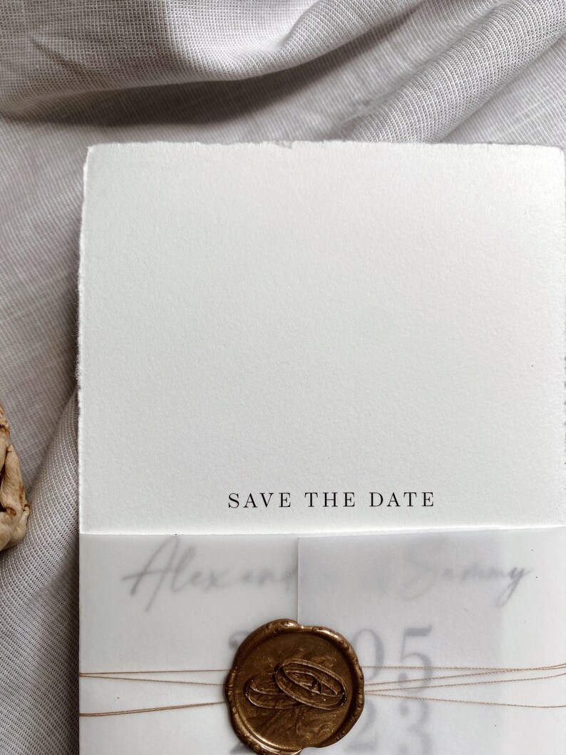 Op de afbeelding: Een witte save-the-date kaart met een gouden waszegel en de tekst "Save the Date" gedrukt op de voorkant. De kaart is vastgebonden met een dun bruin touwtje.