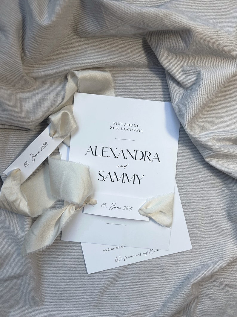 May include: A white wedding invitation with black text. The invitation reads "Einladung zur Hochzeit" which translates to "Invitation to the Wedding". The names Alexandra and Sammy are printed in large, bold letters. The date "08. June 2024" is printed on the invitation and on a separate tag.