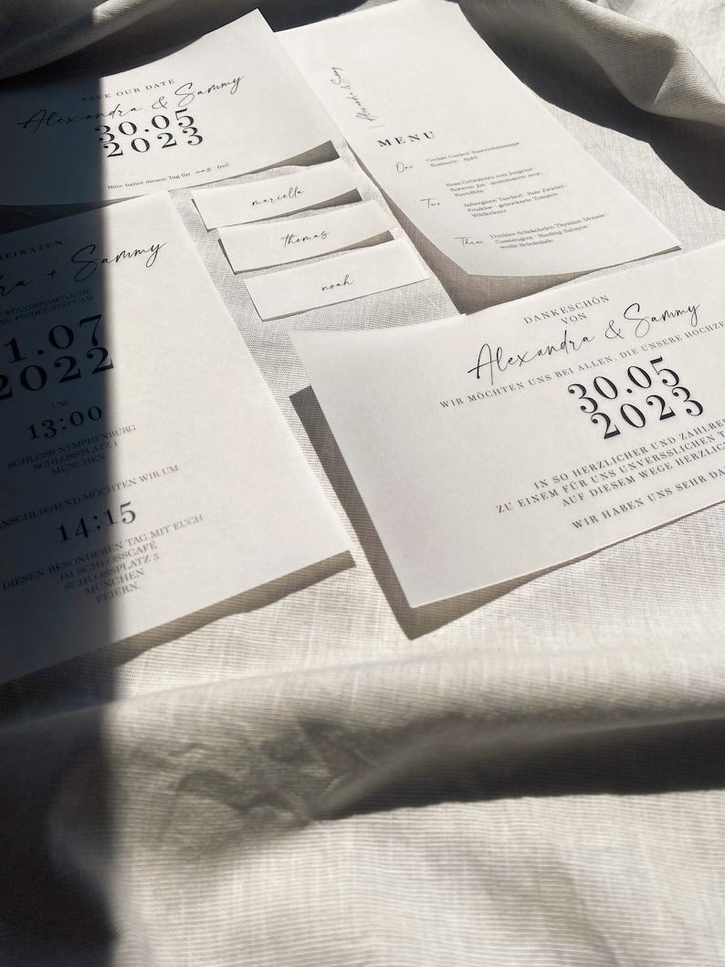 Może przedstawiać: Zestaw zaproszeń ślubnych z białego kartonu z czarną kaligrafią. Zaproszenie zawiera datę "30.05.2023" i imiona "Alexandra & Sammy".