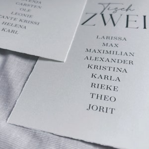 May include: Two white cards with black text. The first card says "Tisch Sieben" and lists names. The second card says "Tisch Zwei" and lists names.
