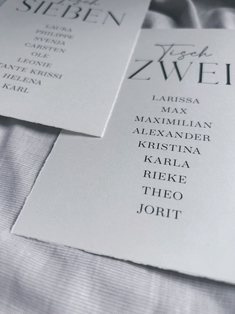 May include: Two white cards with black text. The first card says "Tisch Sieben" and lists names. The second card says "Tisch Zwei" and lists names.