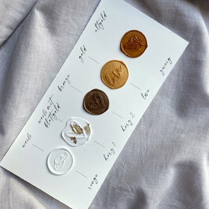 Op de afbeelding: Een witte kaart met waszegels in verschillende kleuren en ontwerpen. De zegels zijn gelabeld met de woorden "wei&szlig;", "blattgold", "wees met bronze", "gold", "altgold", "love", "ringe", "herz 1", "herz 2", "lave" en "zweig".