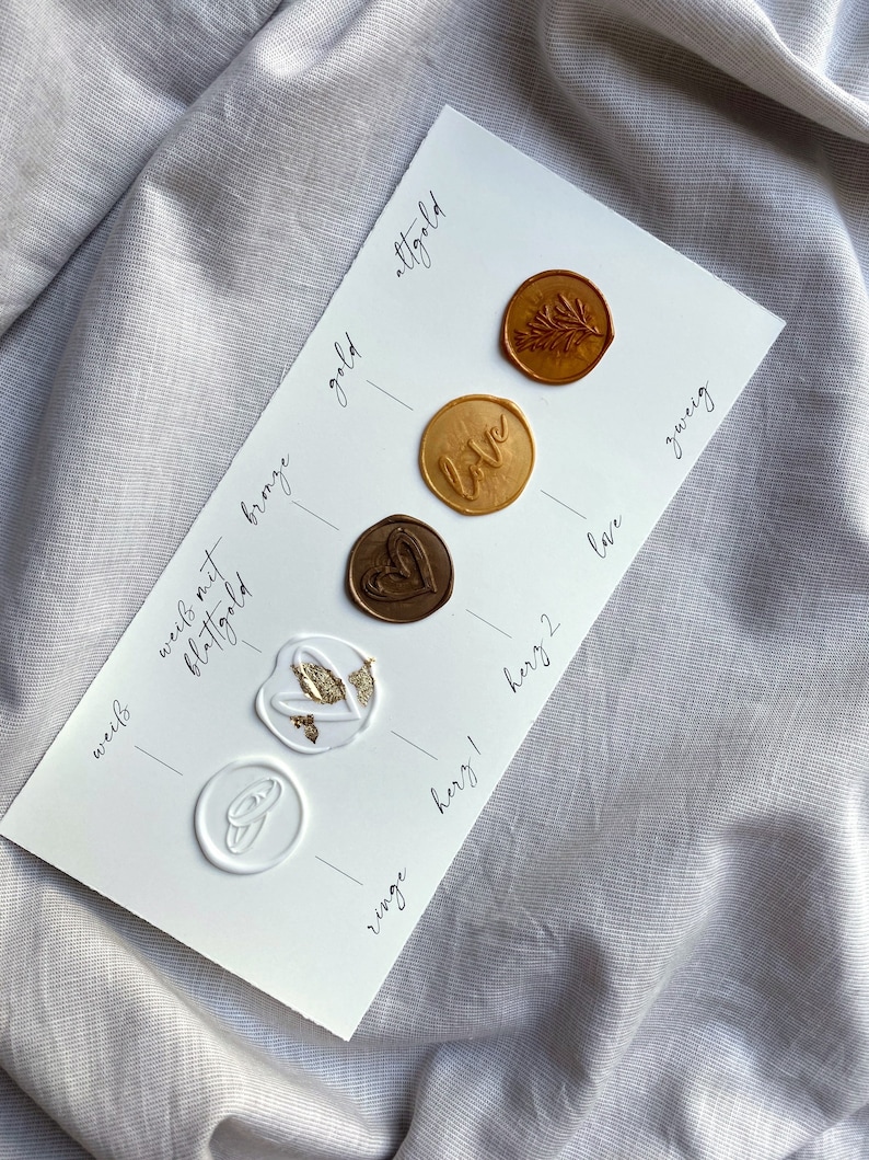 Op de afbeelding: Een witte kaart met waszegels in verschillende kleuren en ontwerpen. De zegels zijn gelabeld met de woorden "wei&szlig;", "blattgold", "wees met bronze", "gold", "altgold", "love", "ringe", "herz 1", "herz 2", "lave" en "zweig".