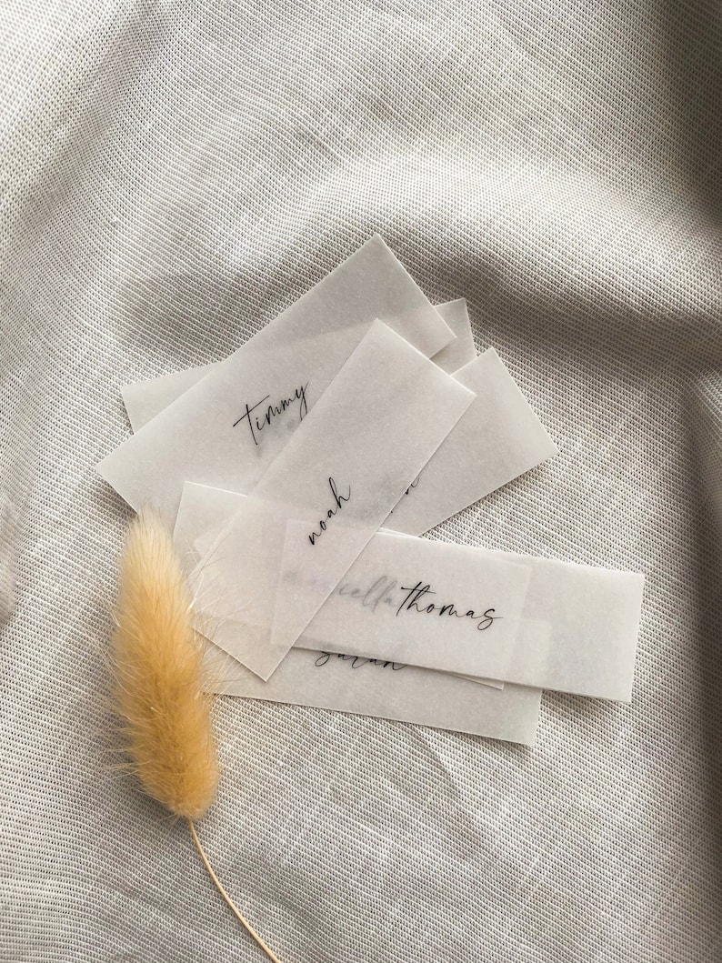 Puede incluir: Una pila de tarjetas de lugar de papel blanco transl&uacute;cido con nombres escritos a mano, incluyendo "Timmy", "Noah", "Kella Thomas" y "Jaren", sobre un fondo de tela texturizada gris claro.