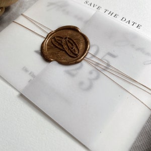 Op de afbeelding: Een witte save-the-date kaart met een gouden waszegel met twee overlappende ringen. De kaart heeft de tekst "Save the Date" erop gedrukt.