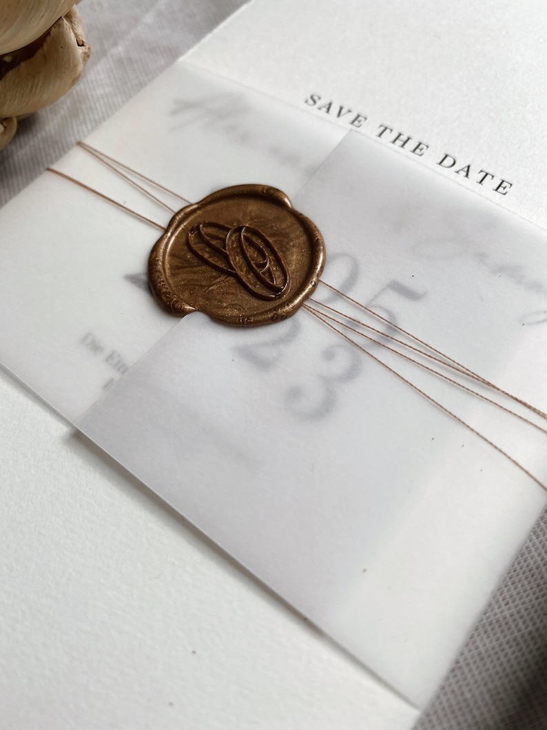 Op de afbeelding: Een witte save-the-date kaart met een gouden waszegel met twee overlappende ringen. De kaart heeft de tekst "Save the Date" erop gedrukt.