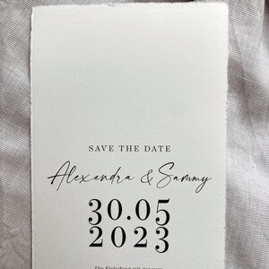 Op de afbeelding: Save the date kaart met een witte achtergrond en zwarte tekst. De tekst luidt "Save the Date" gevolgd door "Alexandra & Sammy" in cursief schrift. De datum "30.05 2023" wordt weergegeven in groot, vetgedrukt lettertype. De tekst "Die Einladung mit genauen Informationen folgt!" staat onderaan de kaart.