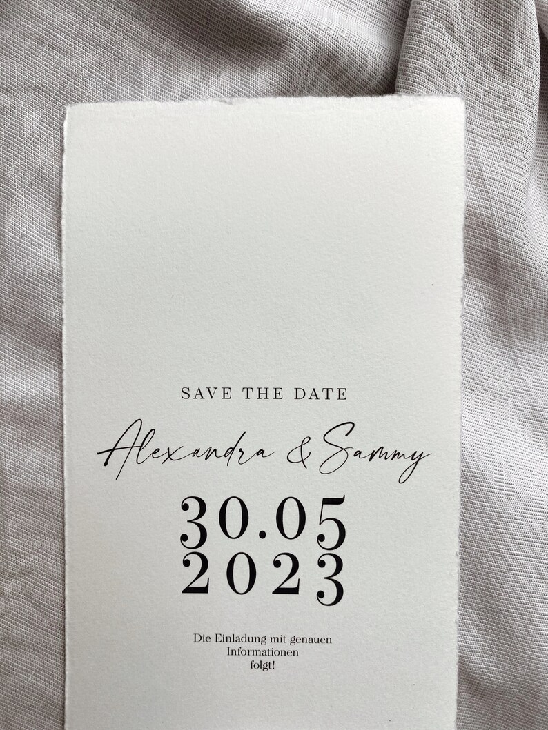 Op de afbeelding: Save the date kaart met een witte achtergrond en zwarte tekst. De tekst luidt "Save the Date" gevolgd door "Alexandra & Sammy" in cursief schrift. De datum "30.05 2023" wordt weergegeven in groot, vetgedrukt lettertype. De tekst "Die Einladung mit genauen Informationen folgt!" staat onderaan de kaart.