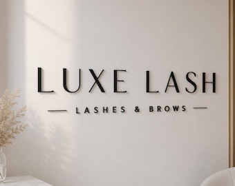 Może przedstawiać: Naścienny szyld z czarnymi, trójwymiarowymi literami tworzącymi napis LUXE LASH, umieszczony nad mniejszym tekstem LASHES & BROWS, otoczonym dwiema poziomymi liniami, wszystko na gładkiej, jasnej powierzchni ściany wewnętrznej.