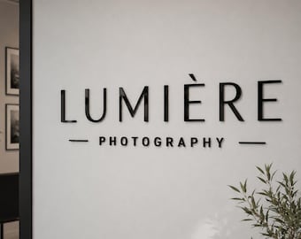Puede incluir: Un letrero de pared con las letras negras tridimensionales "LUMIÈRE" en una fuente sans-serif limpia. Debajo, la palabra "PHOTOGRAPHY" aparece en una fuente negra más pequeña y a juego, flanqueada por dos líneas horizontales negras.