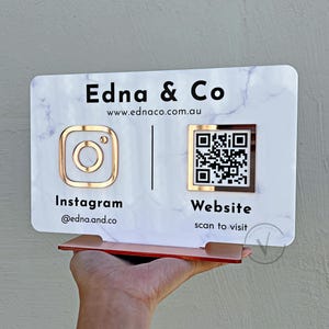 May include: A rectangular sign with a marble effect, displaying "Edna & Co" and a website address. It features a gold Instagram logo and a QR code for the website. The sign is supported by a copper-coloured stand.